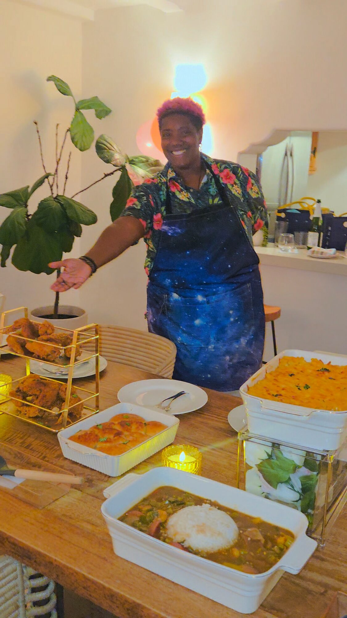 Person with short hair and colorful clothing standing behind a table with food containers, smiling, in a room with a plant and kitchen items.