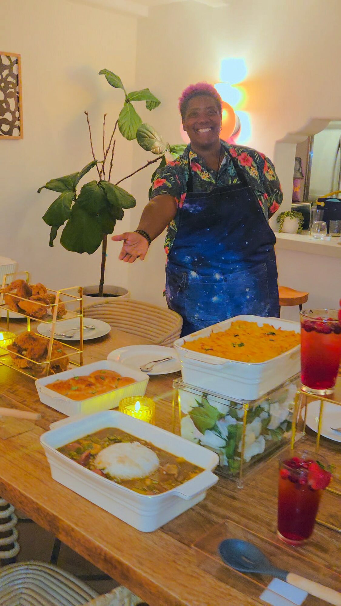 Person smiling in a colorful shirt and apron standing behind a table with food dishes, in a cozy room with a plant and lamp.