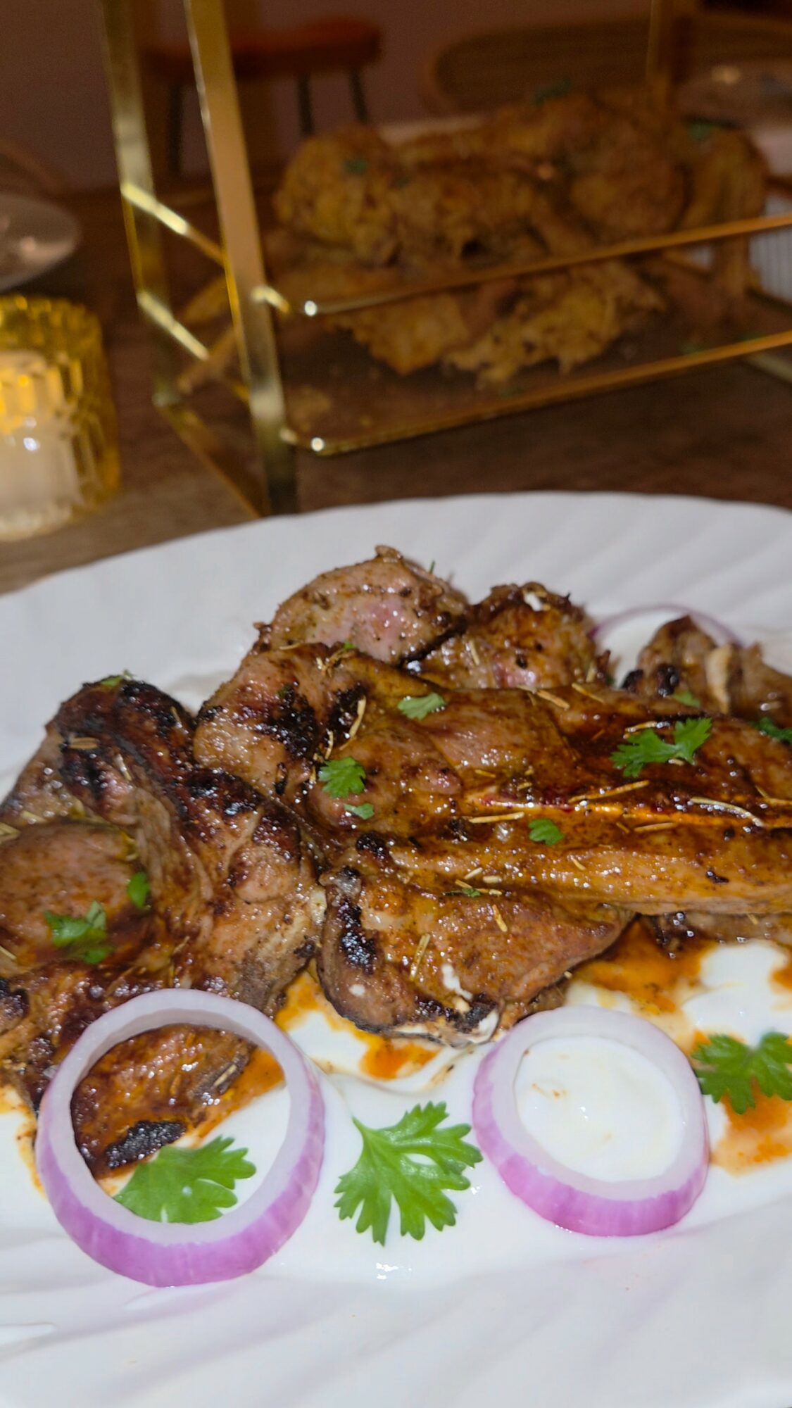 Grilled meat with onion slices and cilantro on a white plate, with fried chicken in the background.