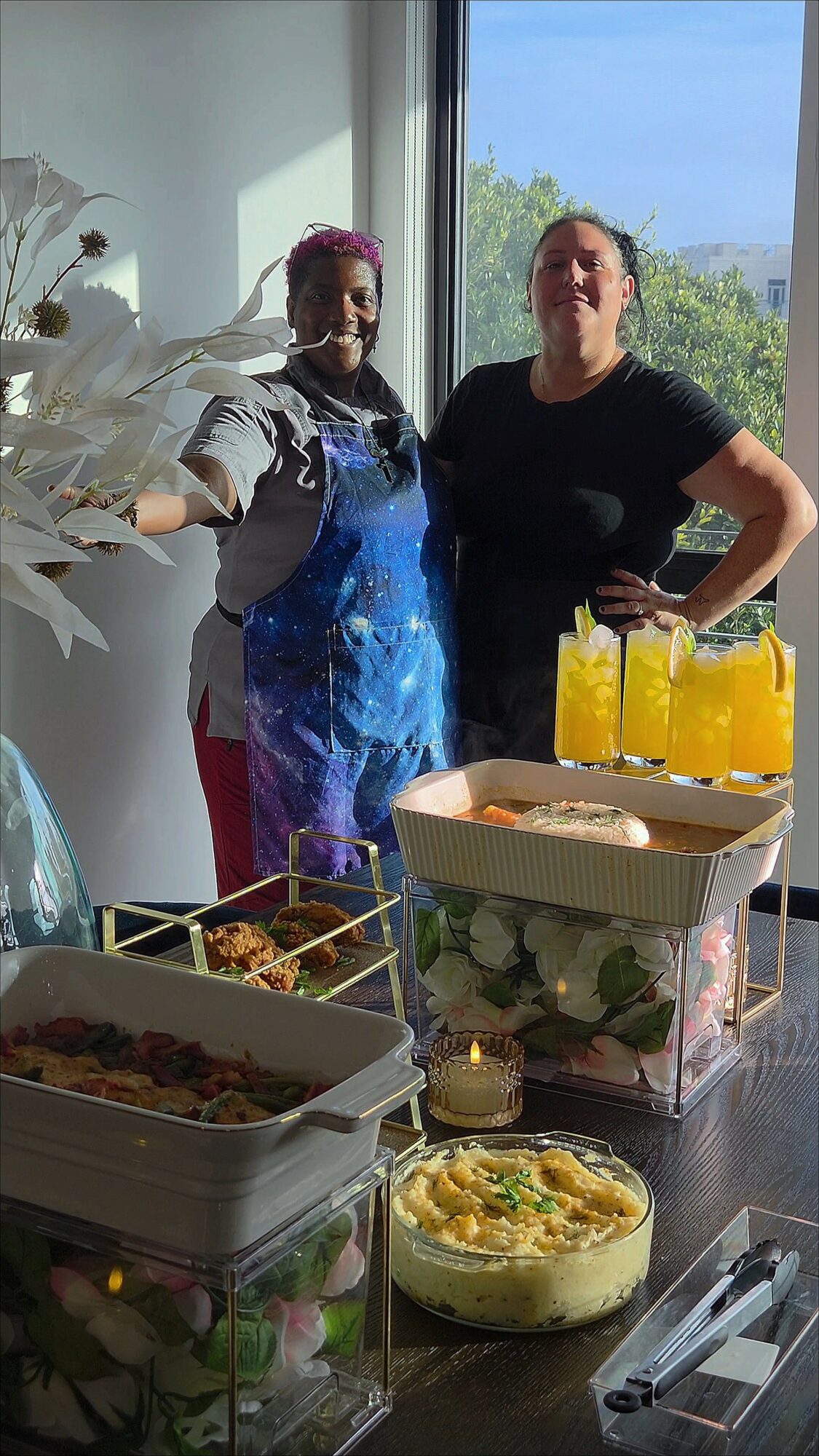 Two women standing behind a table with food and drinks, smiling indoors with a window showing trees outside.
