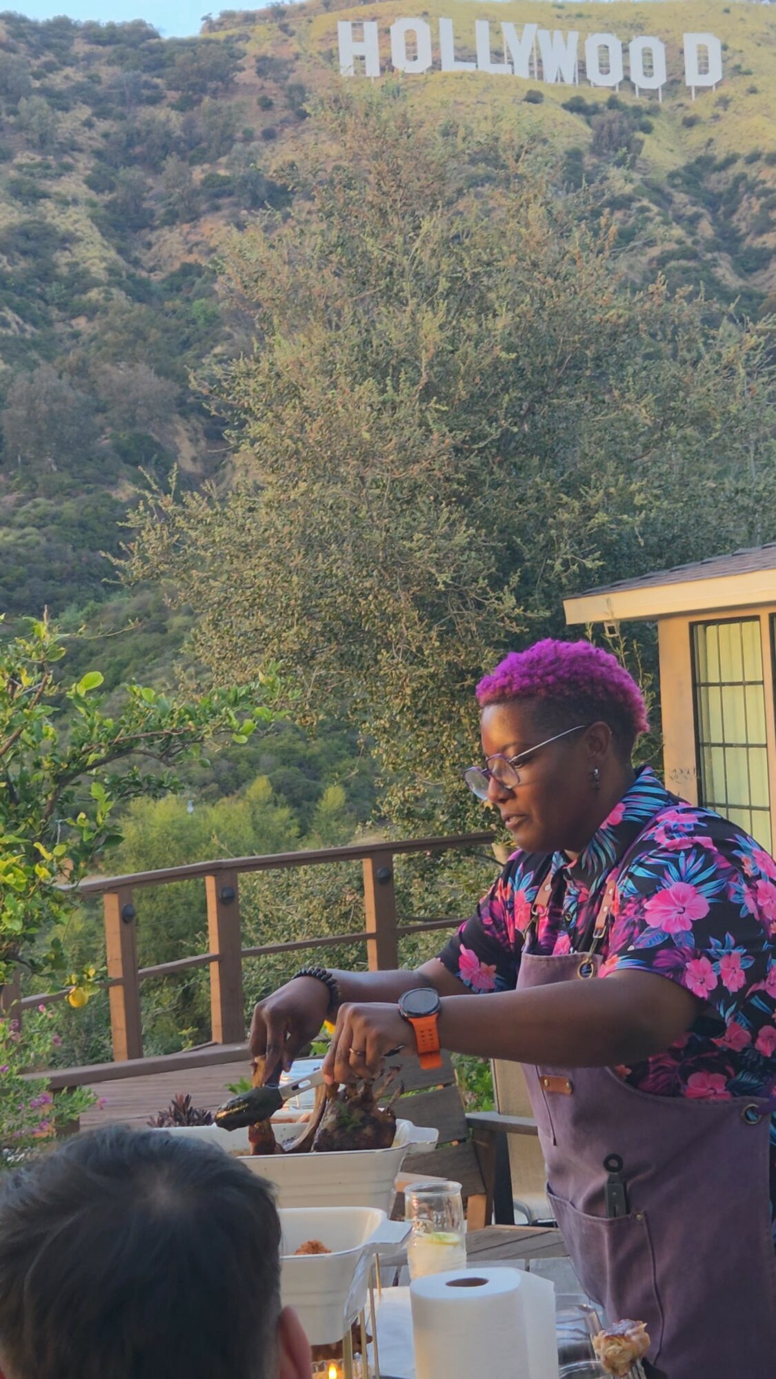 Person with purple hair preparing food outdoors, with Hollywood sign visible in the background.