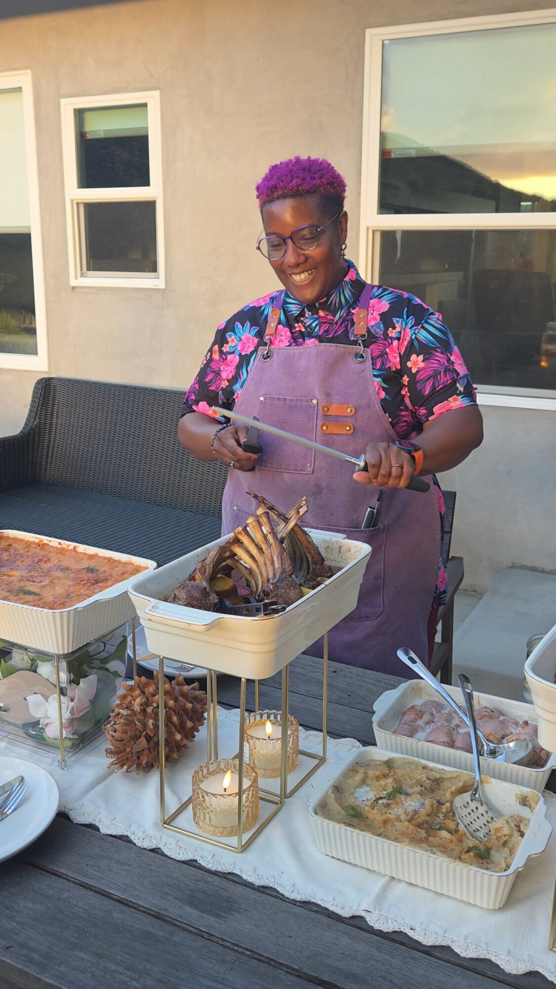 Person with short purple hair wearing a colorful shirt and apron, preparing food outdoors near a house window.