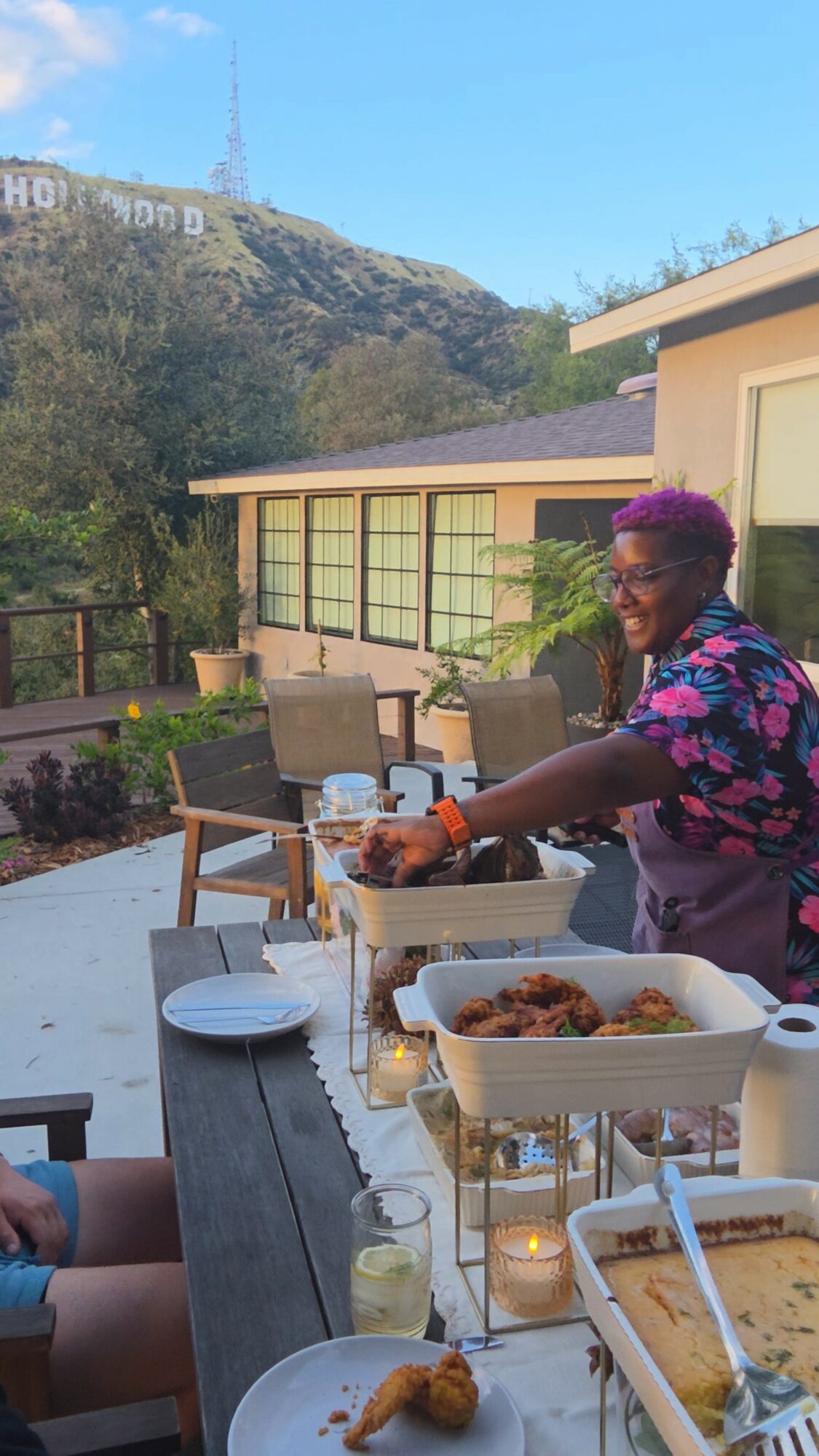 Person with purple hair smiling, serving food outdoors with a hillside and sign in background.