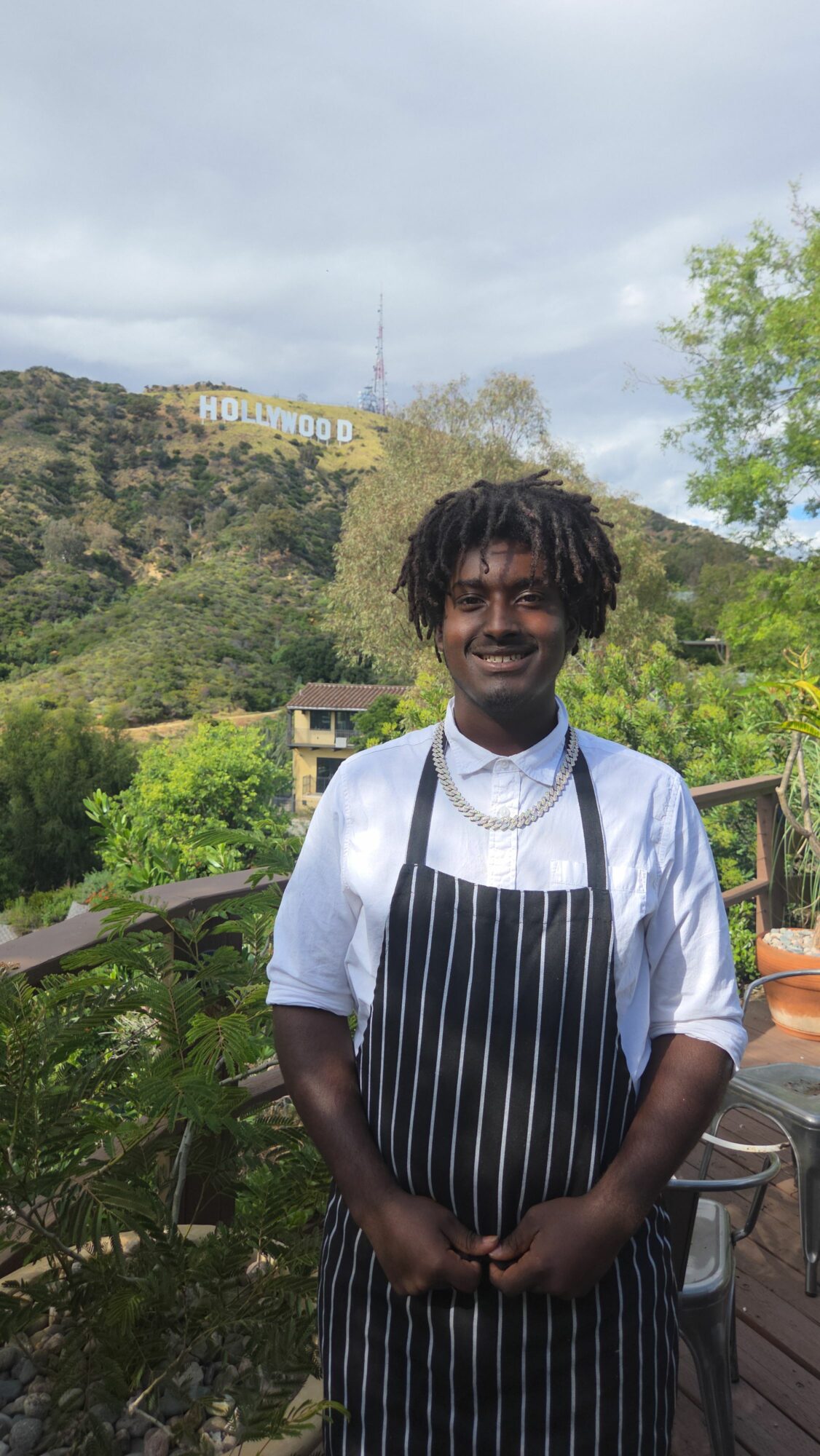 Person with short hair wearing a striped apron over a white shirt, standing outdoors with trees and a hill in the background.