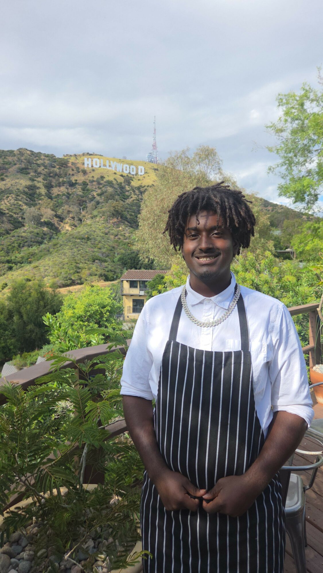 Young person with dreadlocks wearing a striped apron and white shirt outdoors, with trees and a hill in the background.