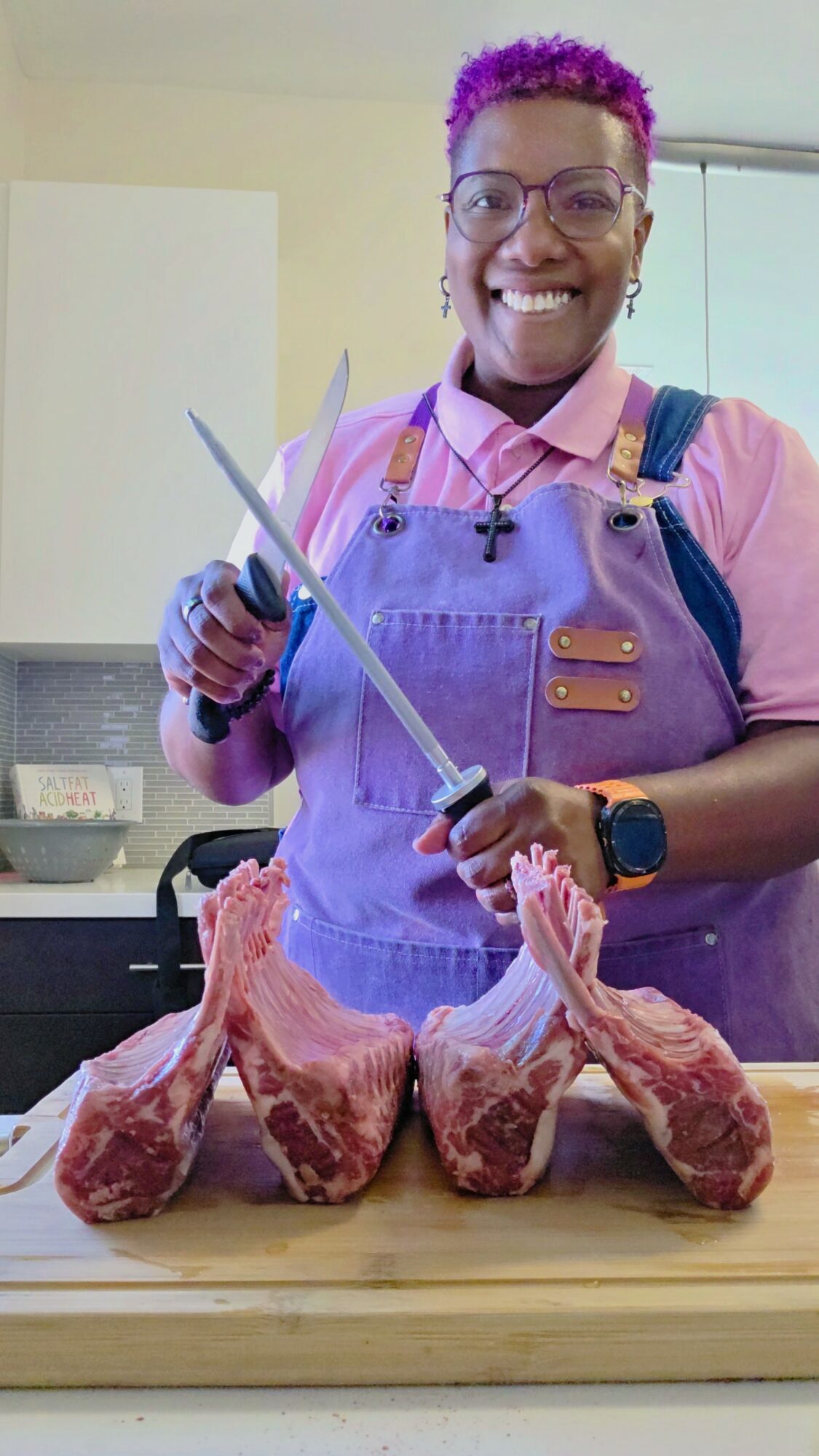 Person with purple hair and glasses smiling, holding a knife, standing behind three raw meat cuts on a wooden surface.