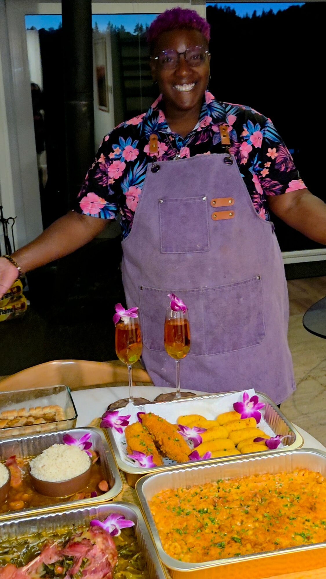 Person with purple hair smiling, wearing a floral shirt and purple apron, standing behind a table with food and drinks.