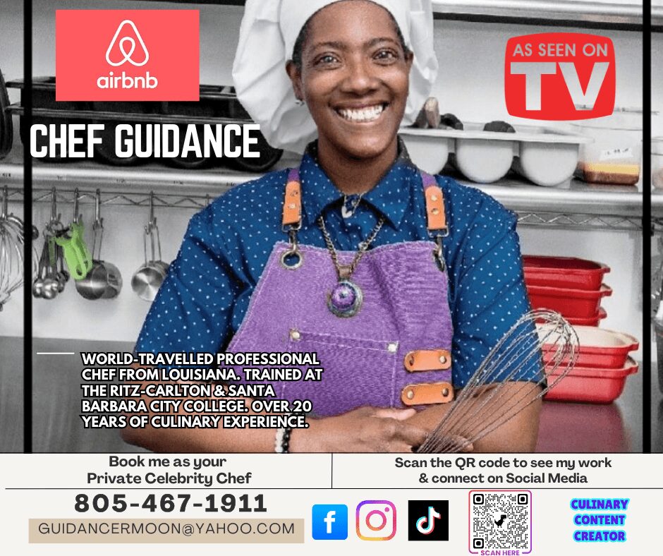 Smiling woman in a kitchen wearing a purple apron and blue shirt, with kitchen utensils and shelves behind her.