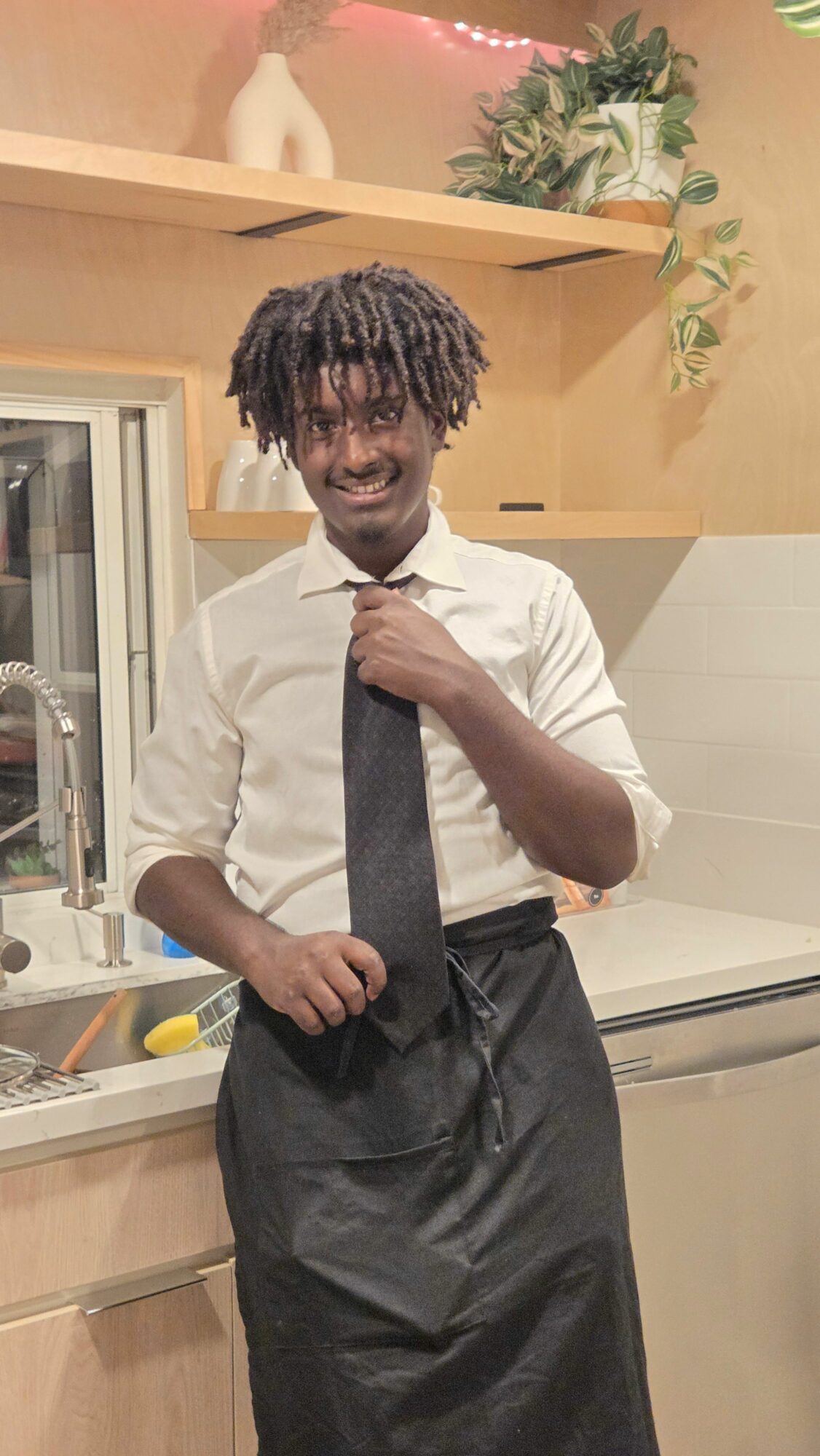 Young person with dreadlocks in a white shirt and black tie, standing indoors near a window and a plant.