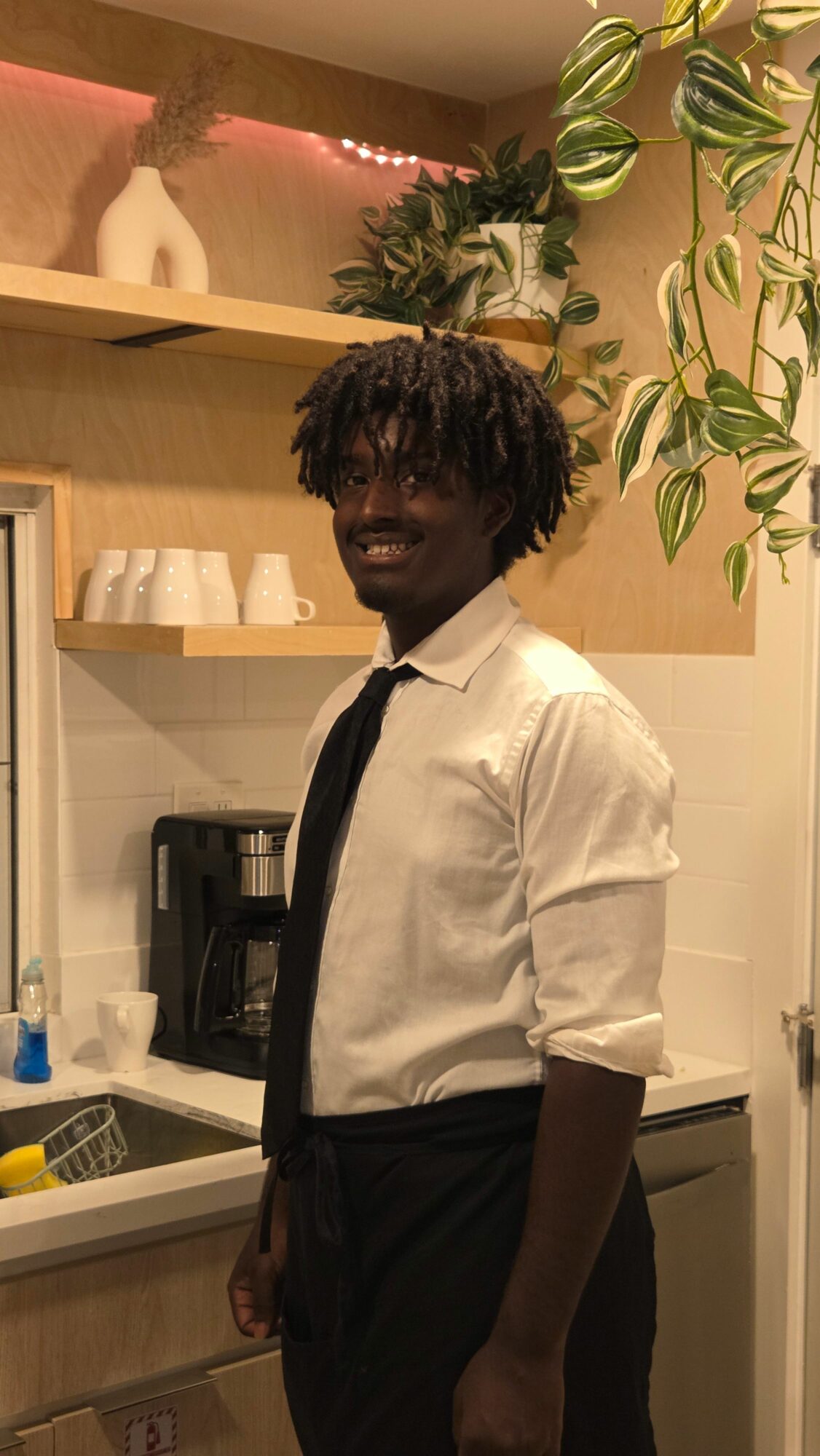 Person with curly hair wearing a white shirt and black pants standing in a room with plants and kitchen items.
