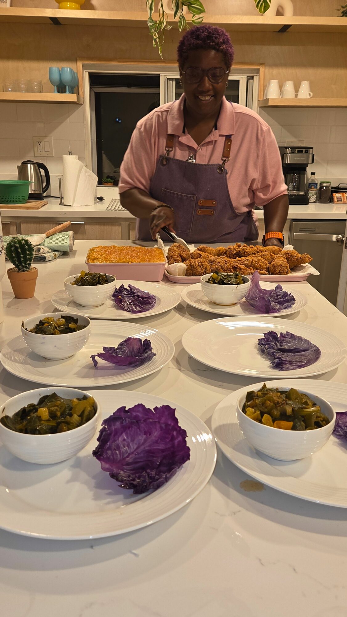 Person in pink shirt preparing food in a kitchen with plates of purple and green dishes on a white table.