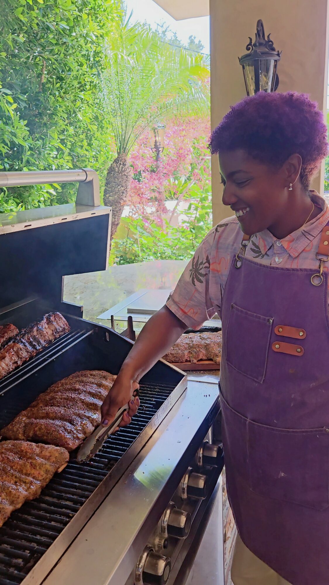 Young woman grilling meat outdoors, smiling, wearing a purple apron, with green trees and pink flowers in background.