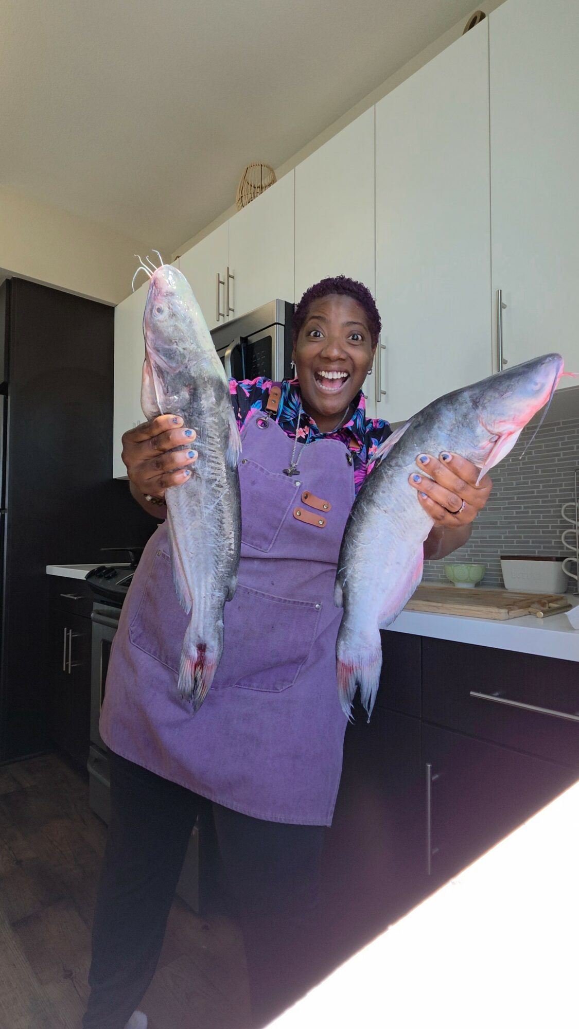 Woman in purple apron holding two large fish in a kitchen, smiling.