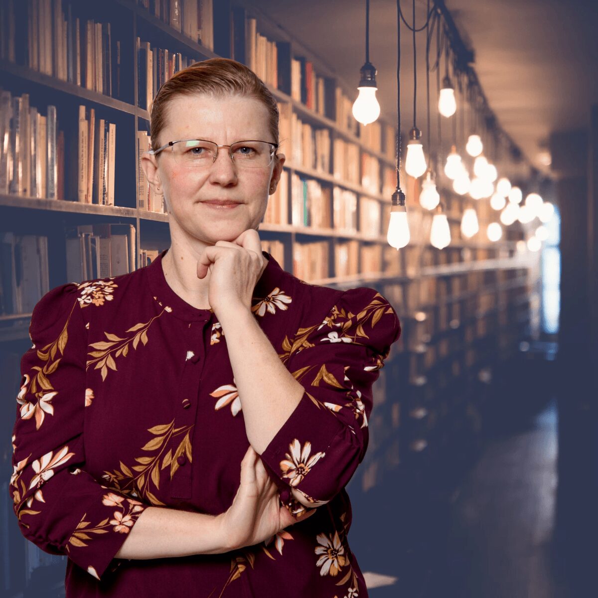 Woman with glasses and short hair, wearing a floral dress, standing in a library with bookshelves and hanging lights behind her.