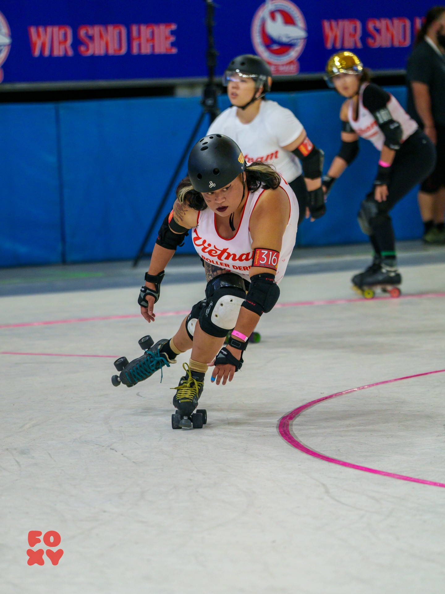 Female roller skater in black helmet and protective gear leans forward on the rink, with three other skaters behind her.