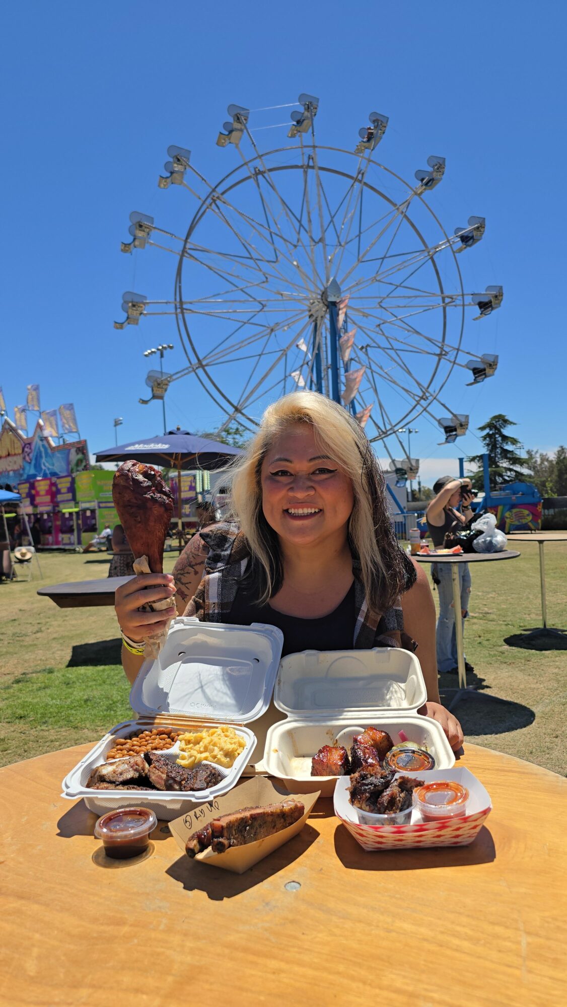 Woman enjoying food at an outdoor fair with a Ferris wheel in the background, smiling and holding a drink.