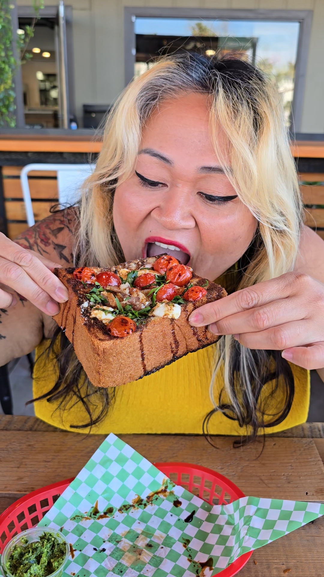 Woman with blonde hair eating a large slice of pizza at a table, with green checkered paper and a red basket in front.