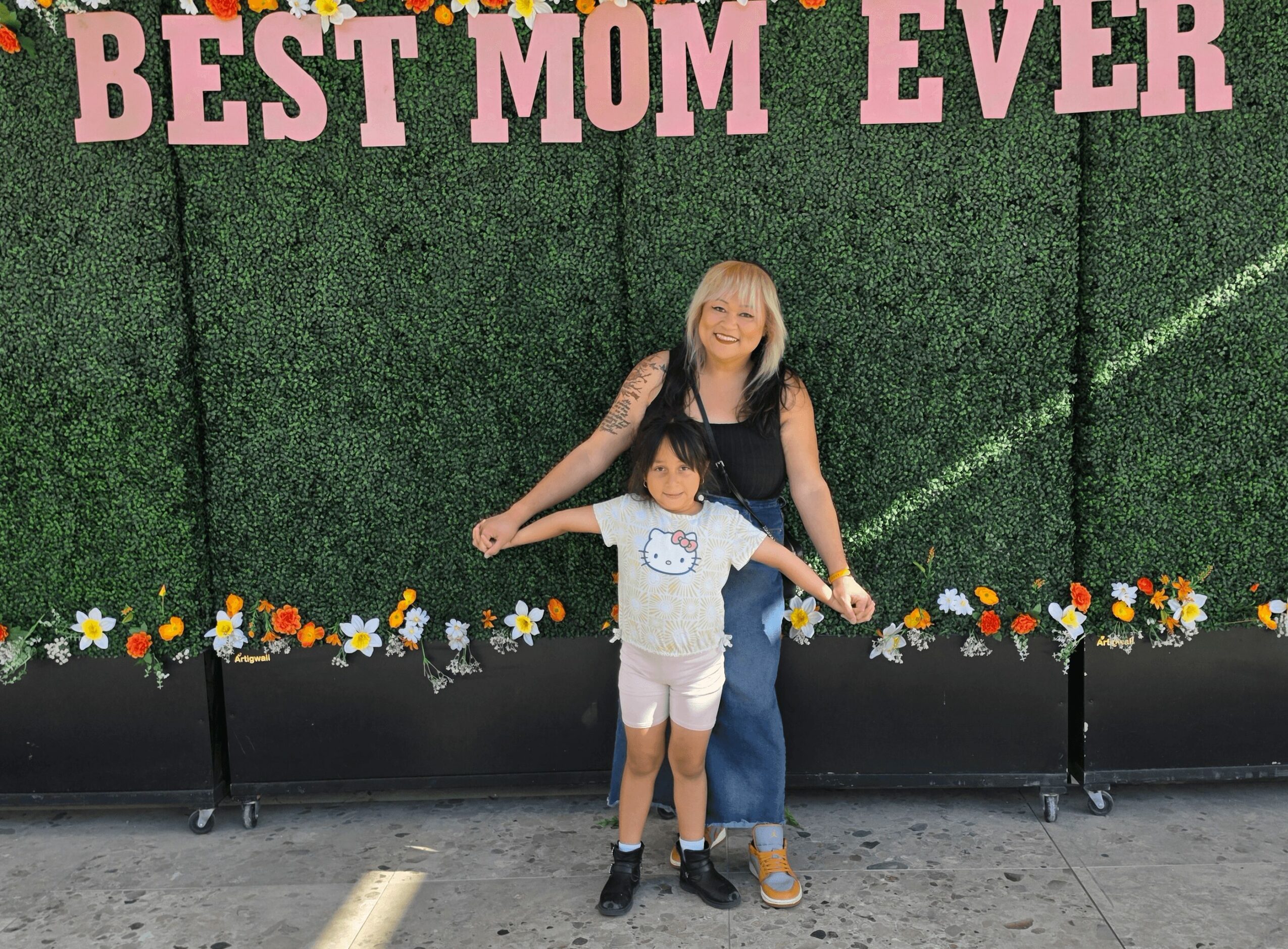 Woman and child standing in front of a green wall with pink letters reading 'Best Mom Ever,' smiling.
