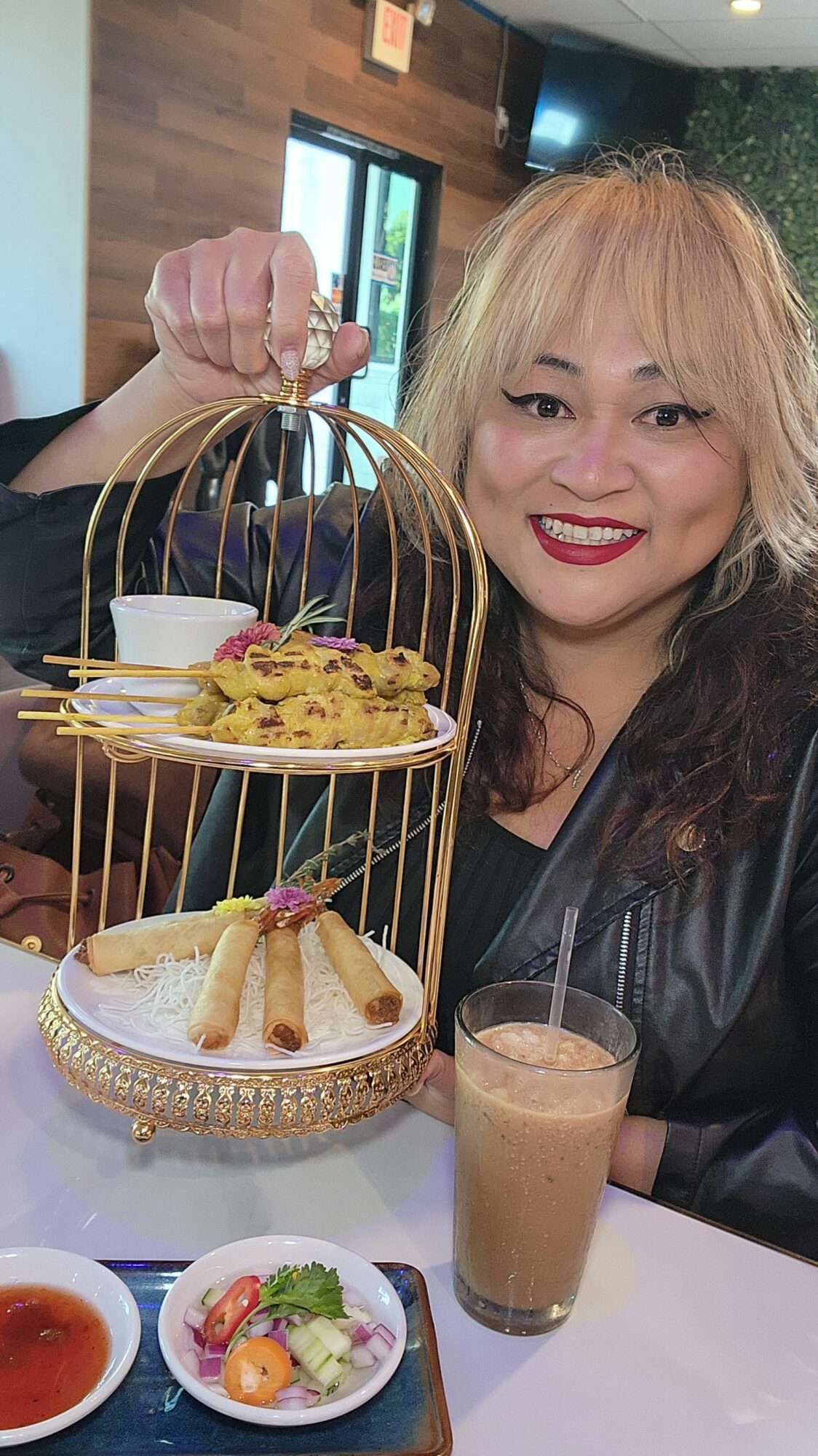 Woman smiling at a table with a multi-tiered dessert stand, coffee, and small plates, in a restaurant setting.