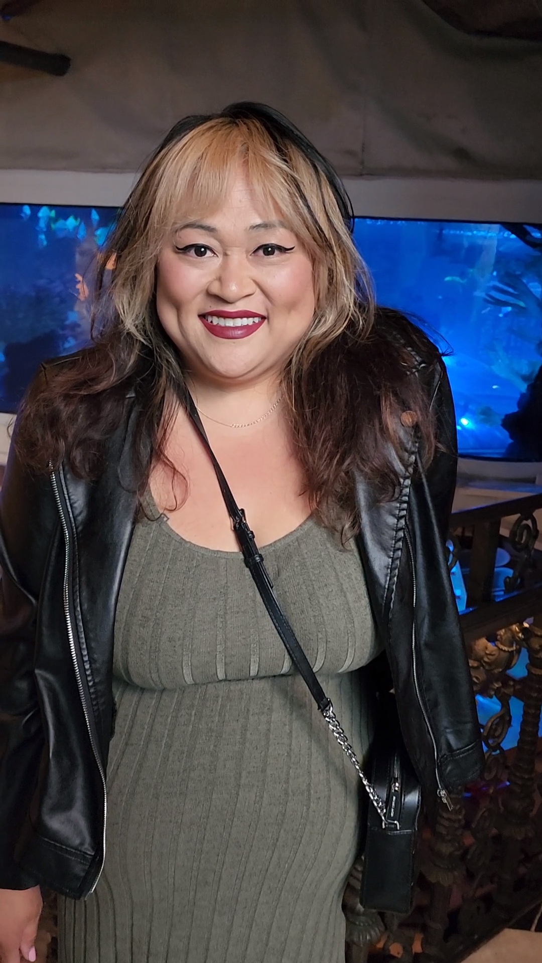 Woman with blonde and dark hair smiling, wearing a green dress and black leather jacket, in front of a blue background.