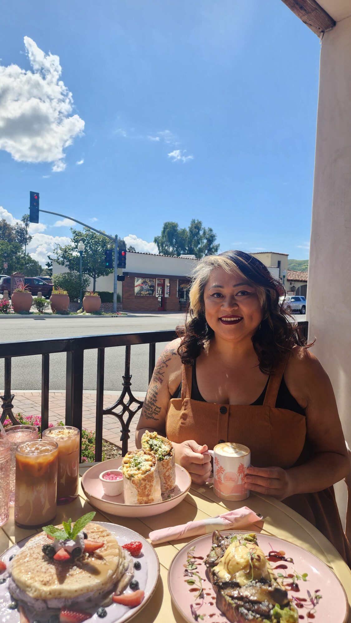 Woman sitting at a table with desserts outdoors, blue sky with clouds in background.
