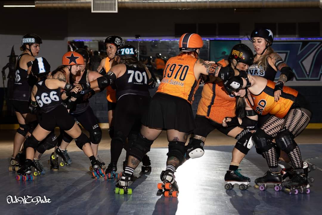 Two teams of roller derby players in black and orange uniforms compete on rink, some blocking and others skating.