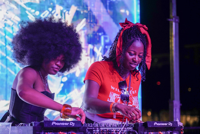Two women with colorful hairstyles and clothing operate DJ equipment on stage with a vibrant background.