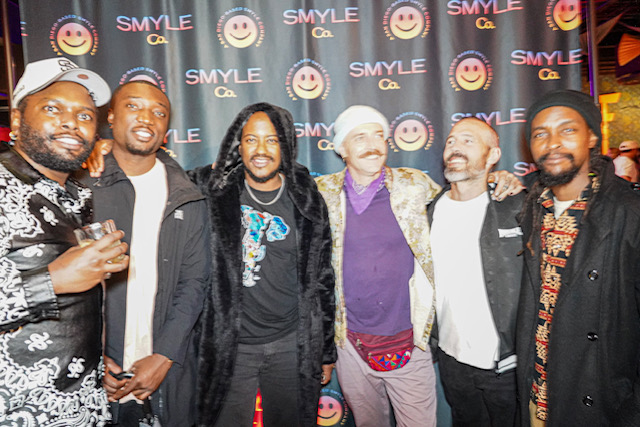 Group of six people smiling and posing in front of a backdrop with smiley faces and the word 'SMYLE'.