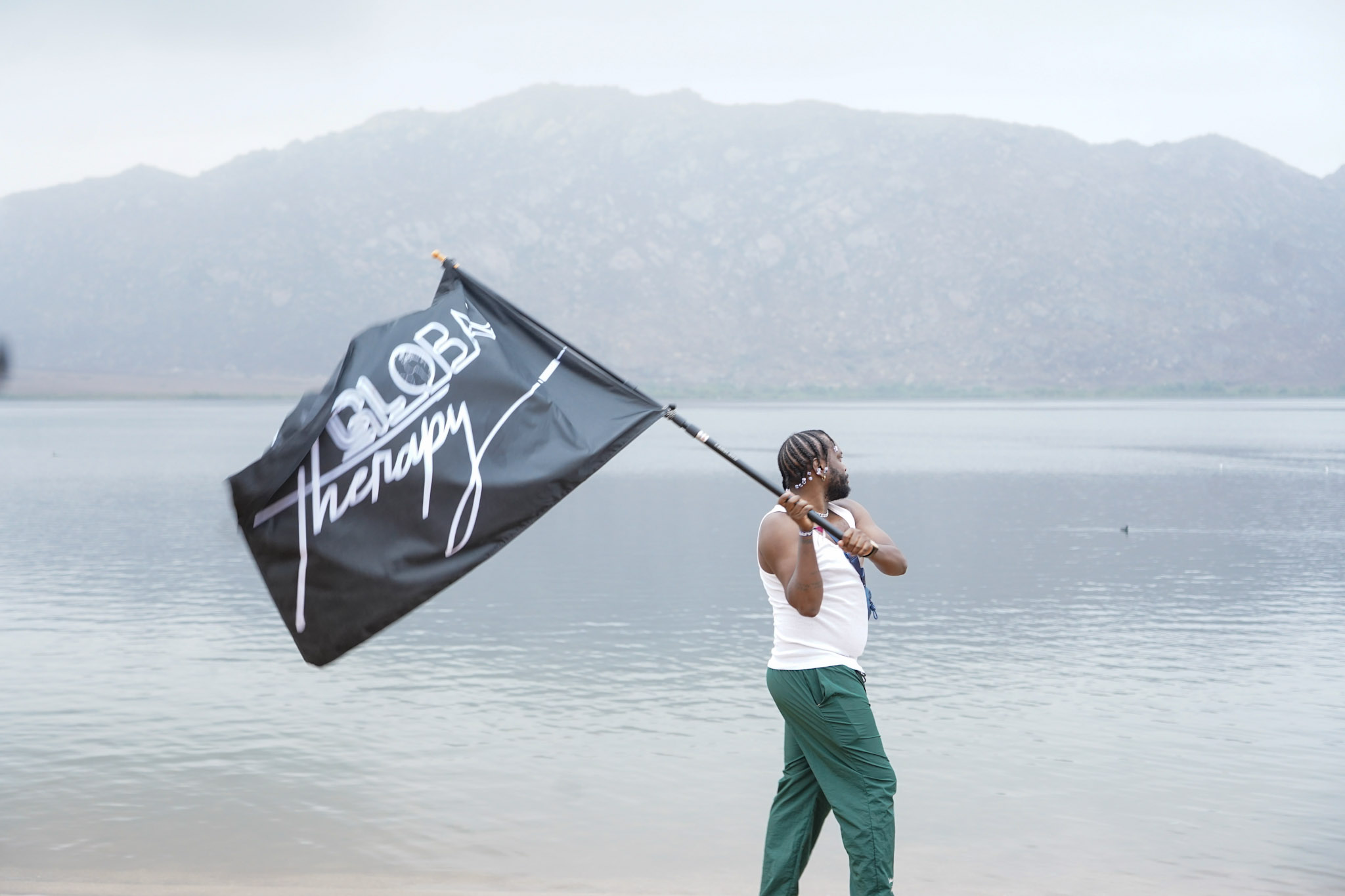 Person standing by water holding a flag with mountains in the background, wearing a white shirt and green pants.