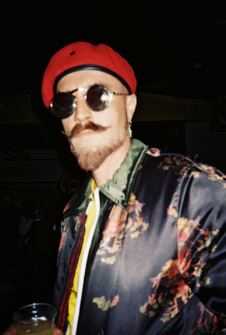 Person with a beard wearing sunglasses, a red beret, and a floral jacket in a dark setting.