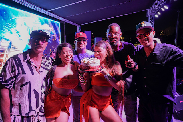 Group of six people celebrating with a cake, smiling, in a dimly lit setting with colorful lights.