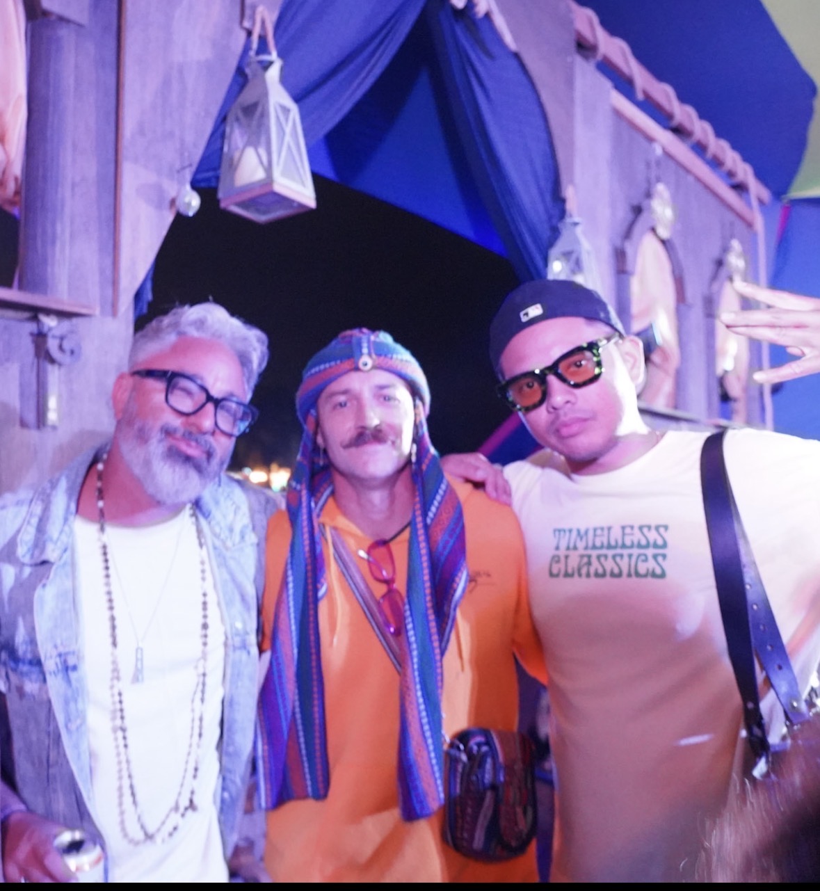 Three people pose together in colorful costumes, with a decorated background and hanging lanterns.