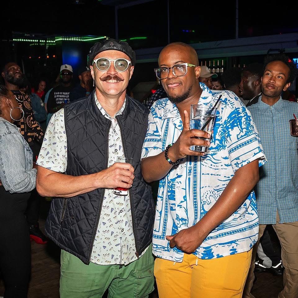 Two men at a party, one with glasses and a mustache, holding a drink, the other bald with glasses, holding a drink, smiling.