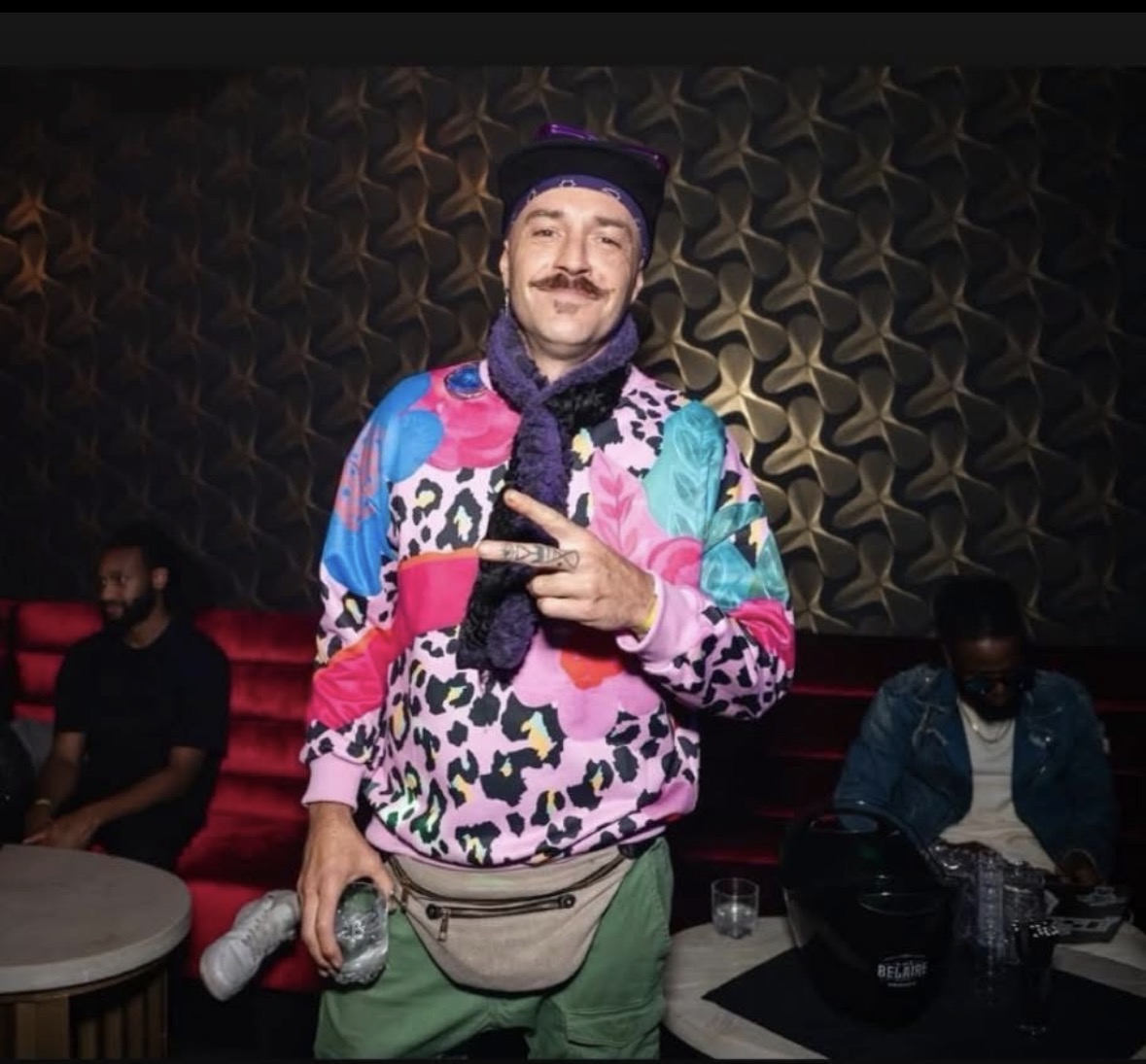 Person with a mustache and colorful patterned jacket, holding a microphone, making a peace sign, in a dimly lit room.