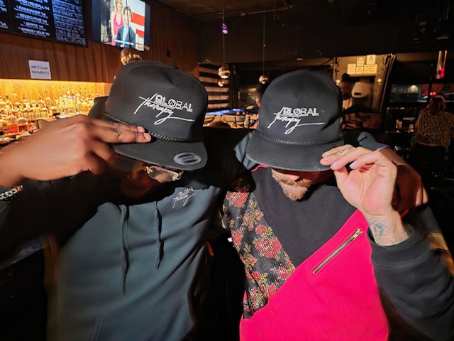 Two people wearing black caps with white text, adjusting their caps in a bar or restaurant setting.