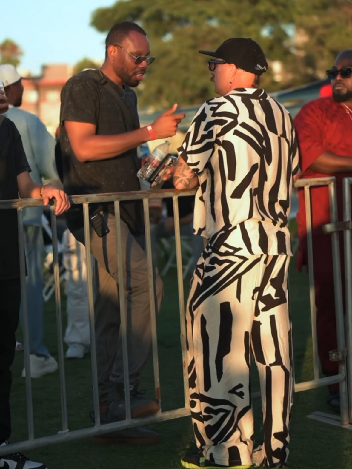 Two men exchanging items outdoors, one in a black shirt and the other in a black and white patterned outfit, with a group nearby.