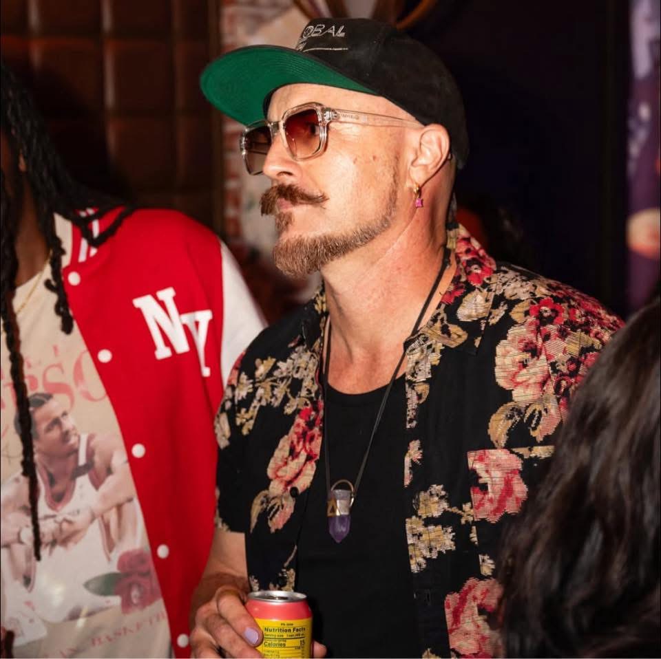 Man with sunglasses, beard, floral shirt, black cap, holding a drink, in a social setting