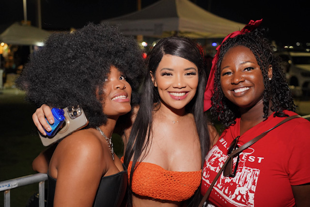 Three women smiling outdoors at night, one with curly hair, one with straight hair, and one with braids wearing a red shirt.