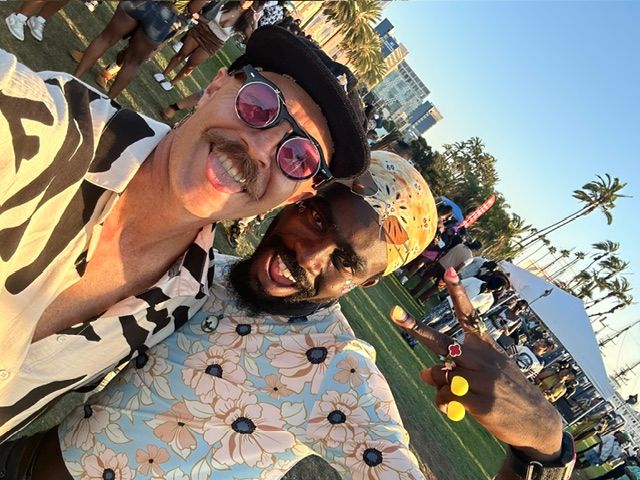 Two people smiling outdoors, one wearing sunglasses and a hat, the other with a patterned shirt, with palm trees and a crowd in the background.