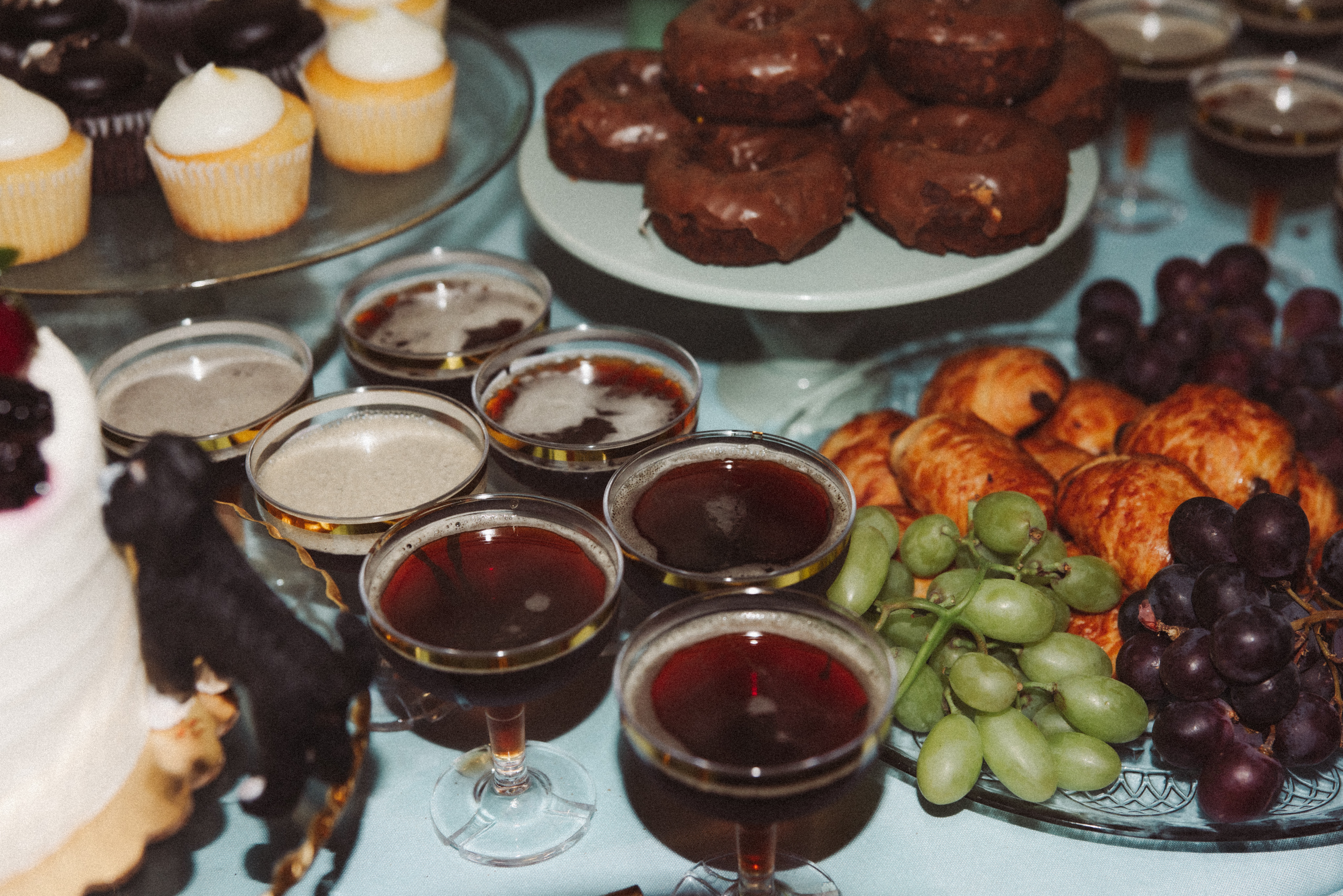 Assorted desserts and fruits on a table, including cupcakes, chocolate muffins, grapes, and small glasses of dark beverages.