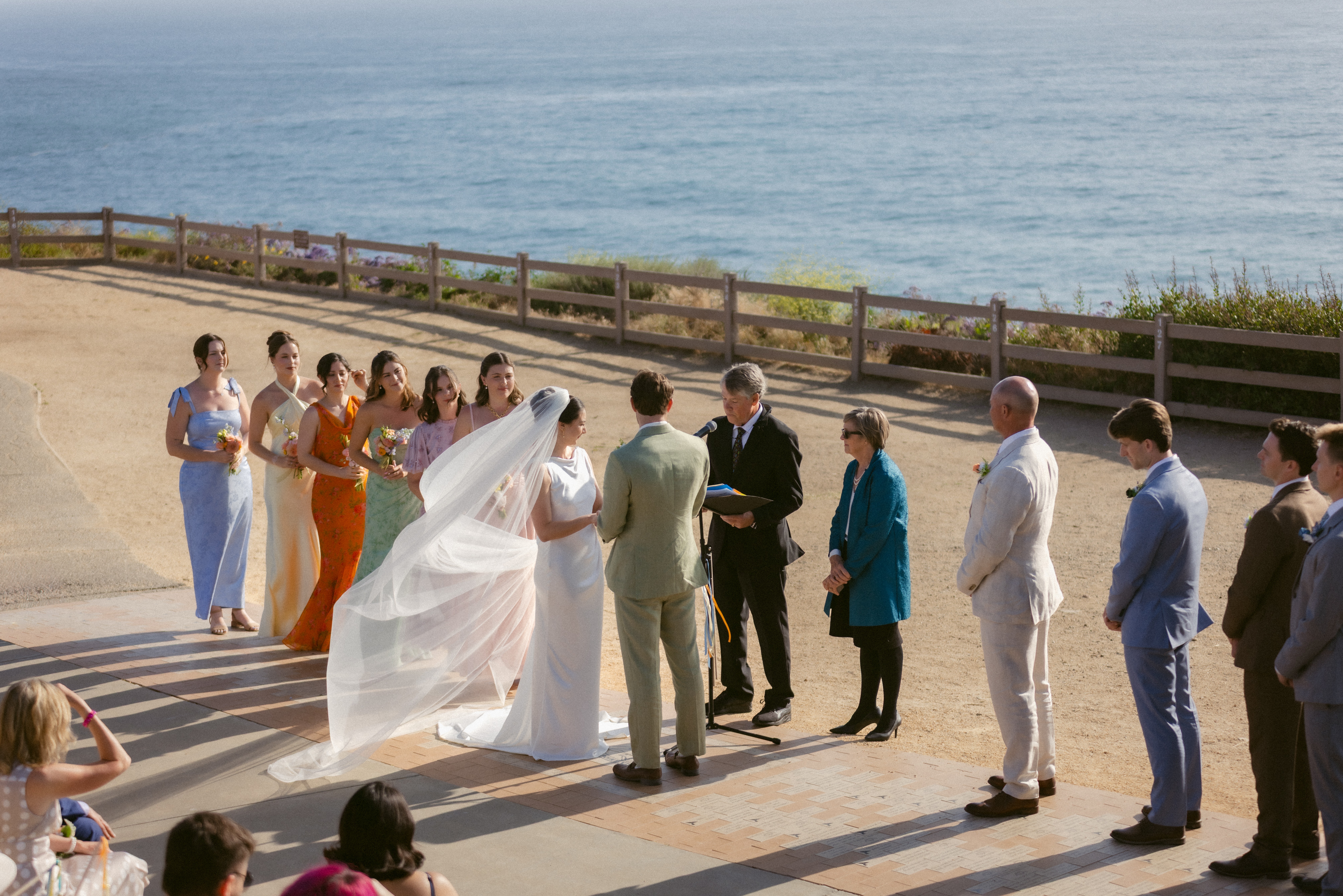 A wedding ceremony outdoors by the water with a bride, groom, officiant, and guests standing in a line.