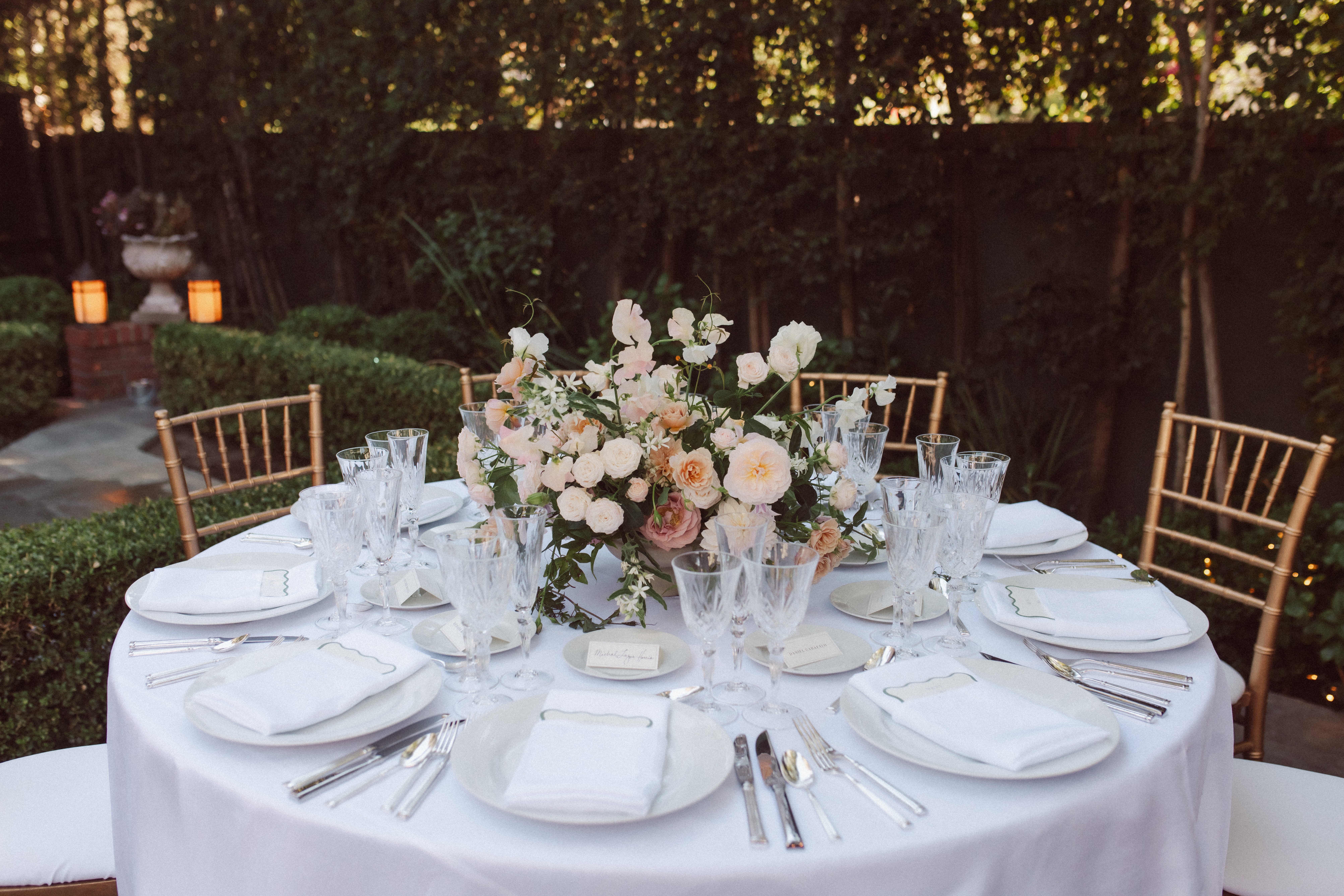 Round table set with white plates, glasses, silverware, and a floral centerpiece outdoors, surrounded by gold chairs.