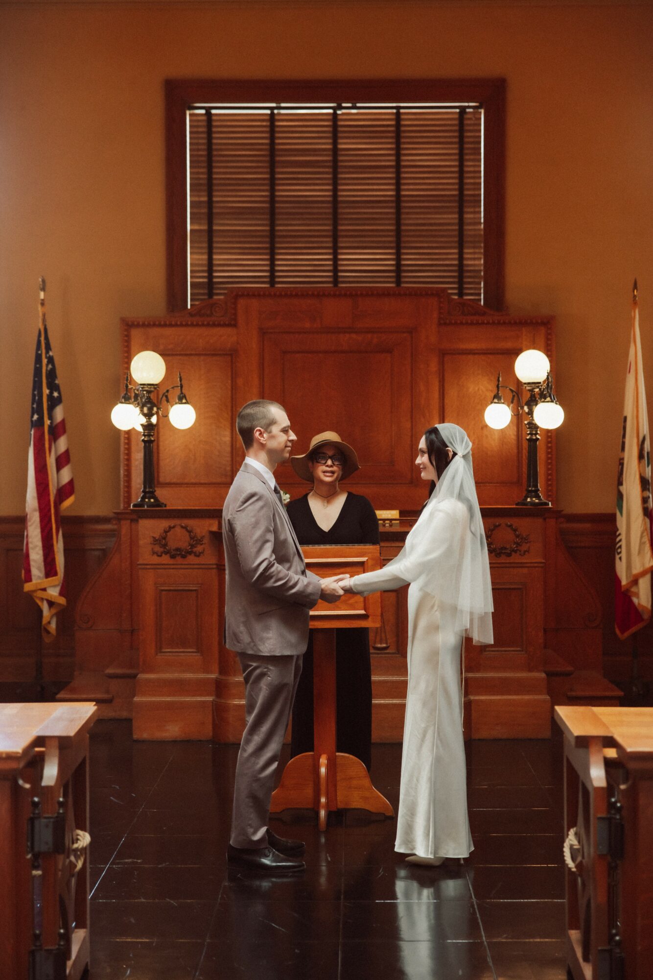 A couple in wedding attire exchanging vows in a courtroom with wooden paneling and American flags, two women officiating, warm lighting.