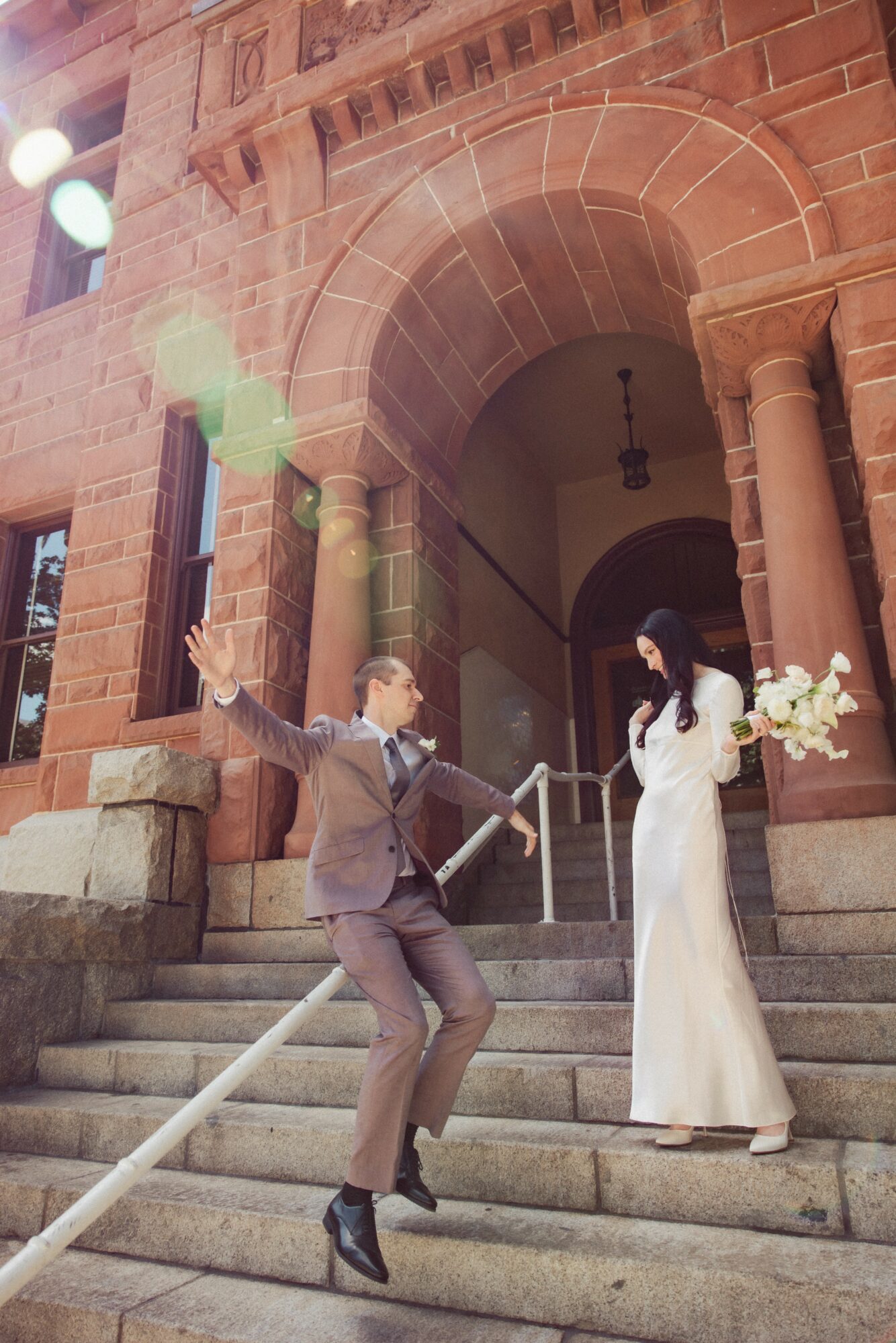 A man in a suit jumps on stairs in front of a woman in a white dress holding a bouquet outside a brick building.