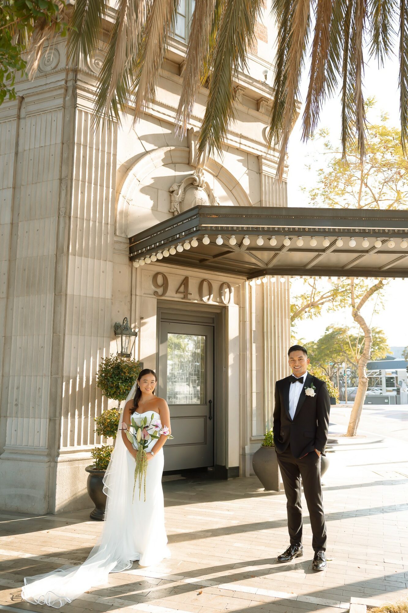 A bride in a white wedding dress and a groom in a tuxedo stand outside a building with tall columns and an arched entrance.