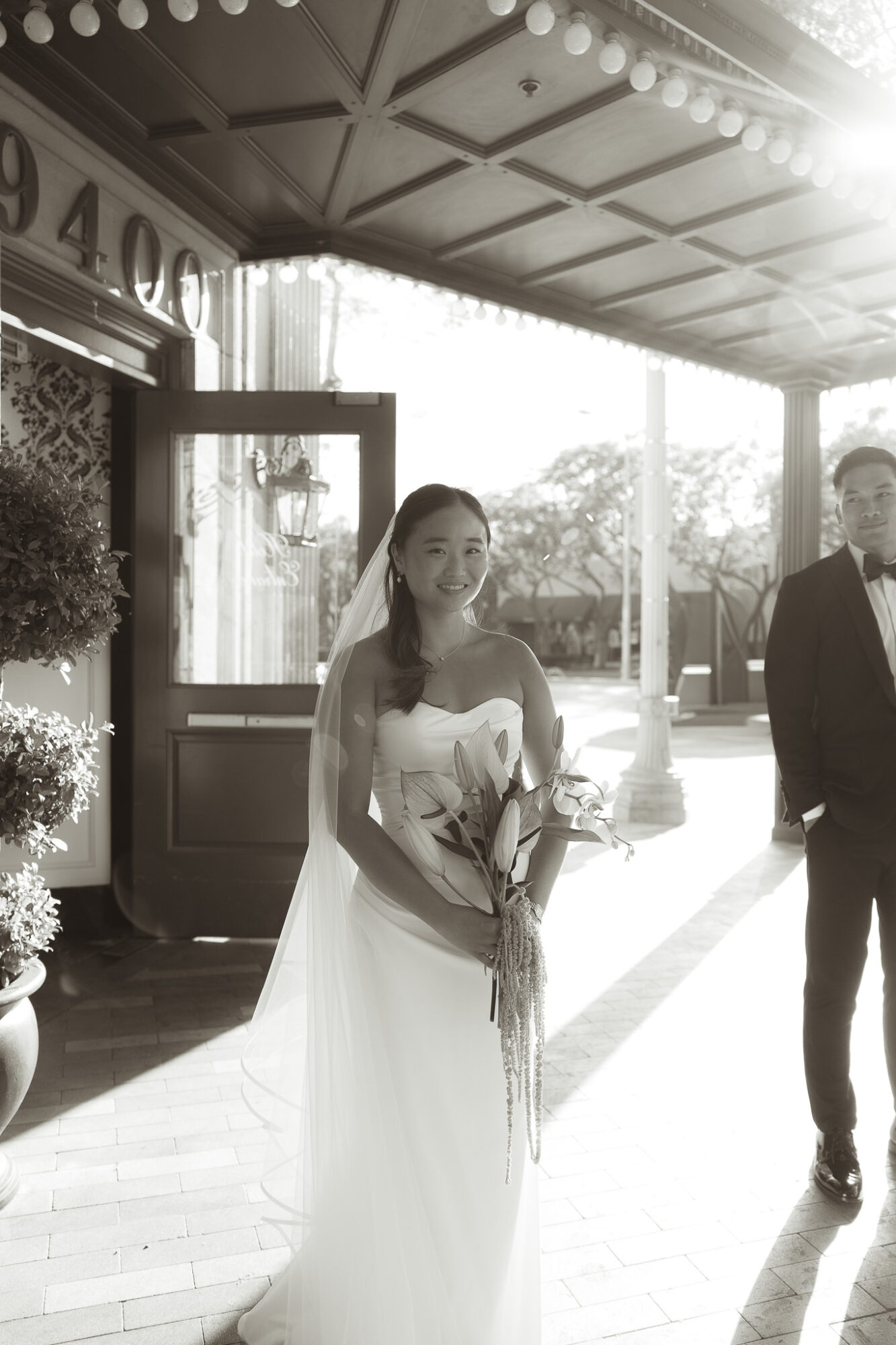 Woman in wedding dress holding flowers, standing outside a building with sunlight, with a man nearby, in black and white.