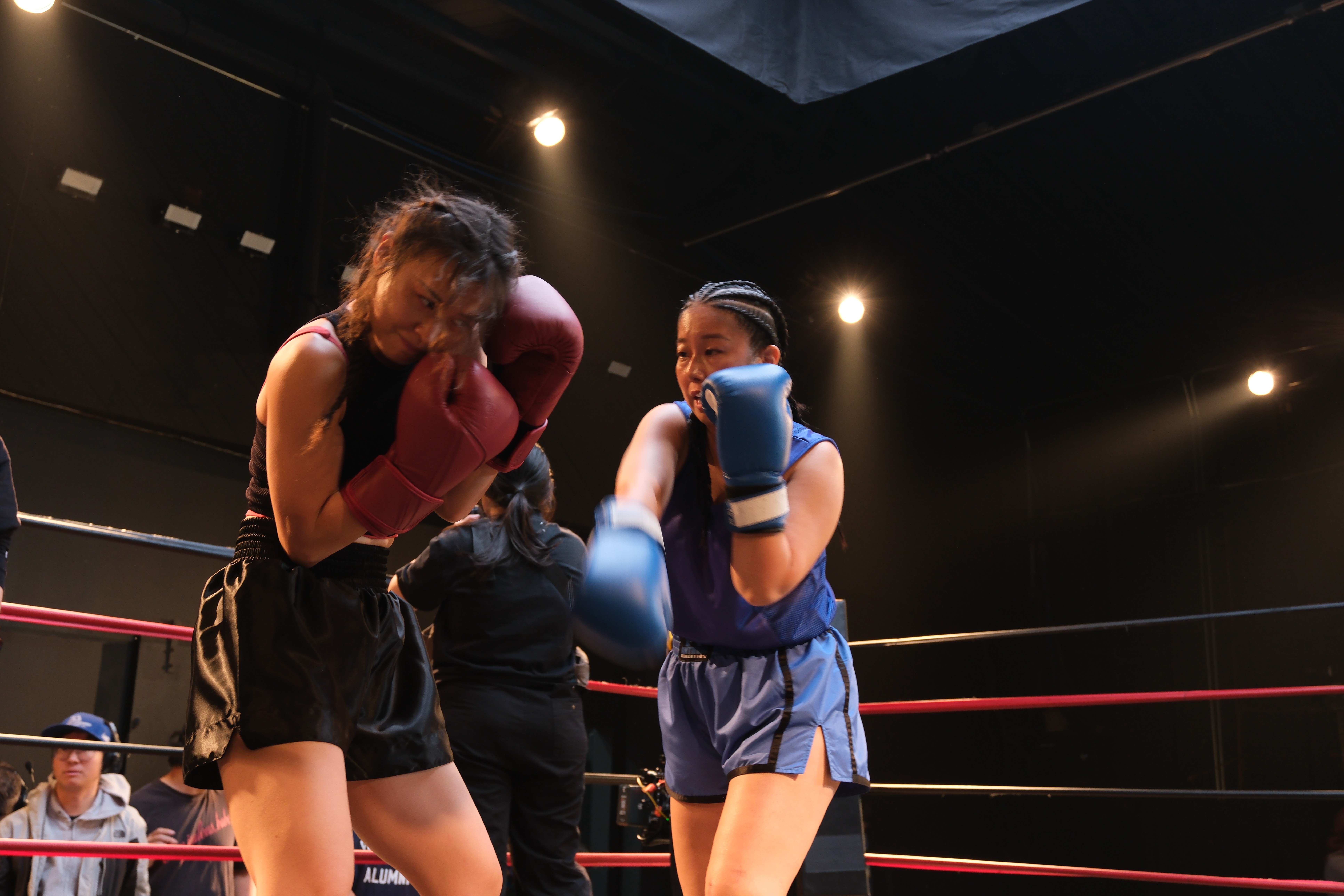 Two young girls in boxing gloves sparring in a boxing ring, with a referee and audience in the background.