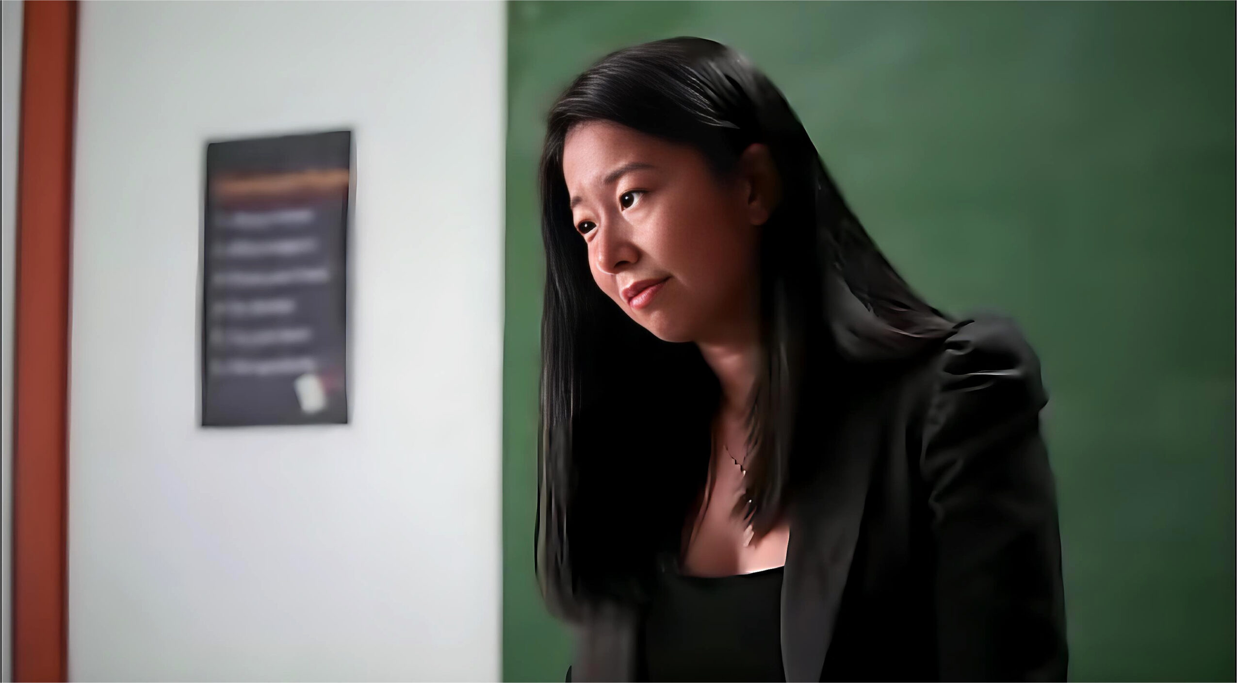 Woman with long black hair looking down in front of a green chalkboard, wearing a black blazer.