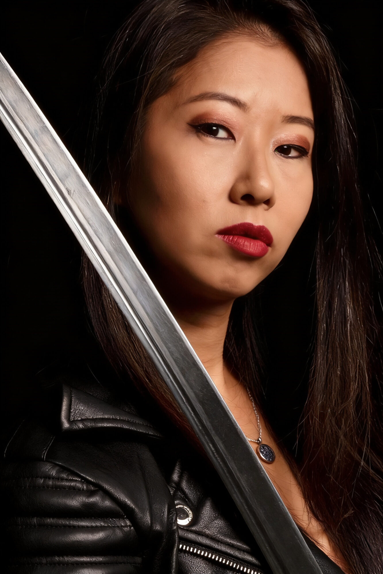 Woman with long dark hair holding a sword, wearing a black leather jacket, looking at the camera.