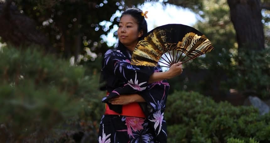 Woman in traditional clothing holding a decorative fan outdoors, surrounded by trees and greenery.